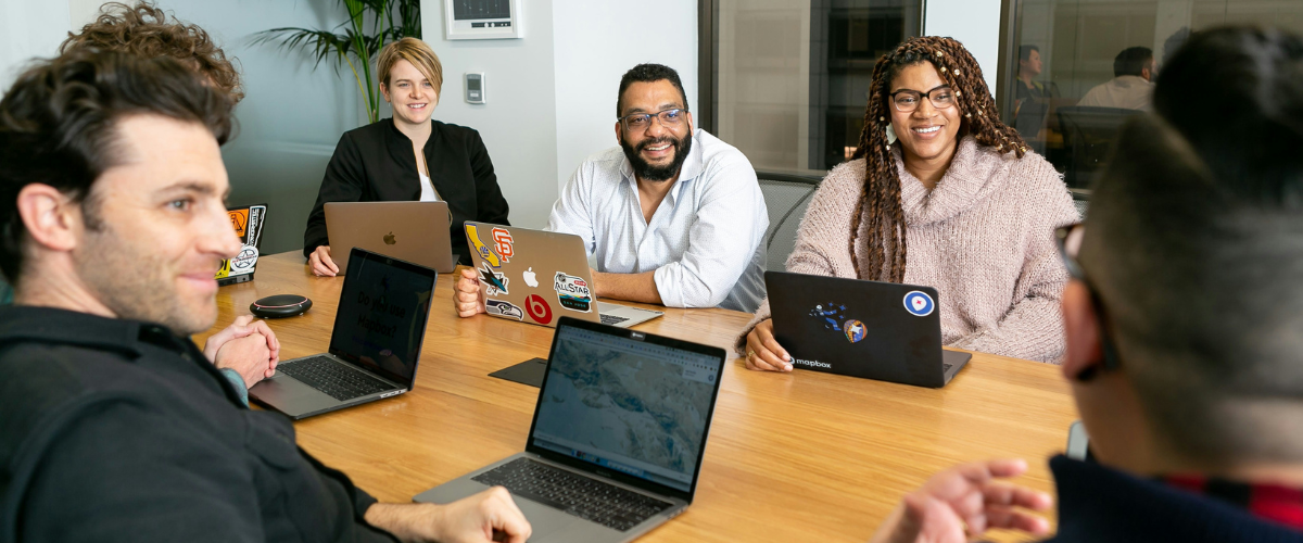 Six colleagues seated around a conference table with open laptops, several smiling toward the camera.